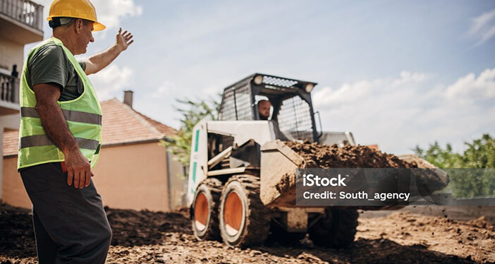 A man in hi-vis and a hard hat assisting a worker using construction equipment to move dirt